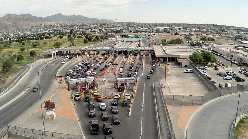 A land border crossing at dusk with lane markings, overhead canopy lights, and a line of waiting cars.