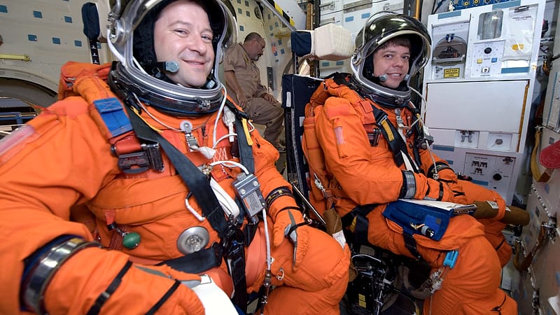 A pressure suit hanging on a rack beside a padded seat and coiled hoses inside a preparation room with bright lighting.