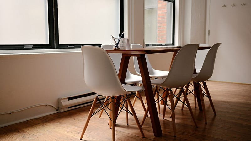 Two upholstered chairs facing each other across a narrow wooden table beside a tall window in an empty room.