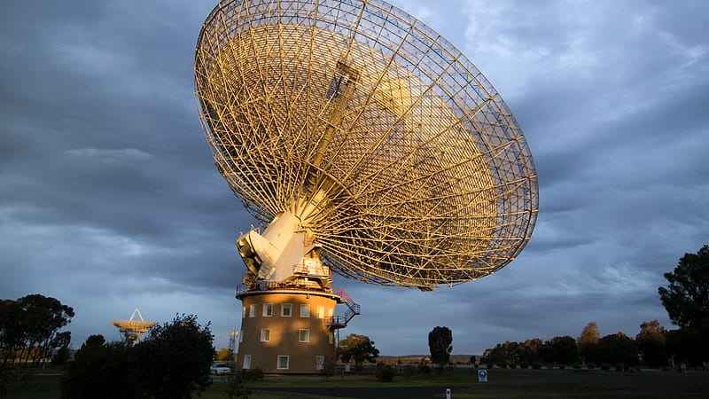 A large steerable dish antenna on a concrete foundation surrounded by a chain link fence with hills in the background.