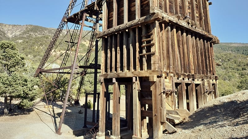 A weathered timber headframe and a corrugated ore mill on a bench of graded rock above a mountain valley.