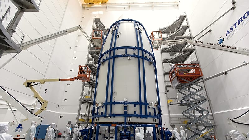 A satellite in a clean room draped in gold foil while two technicians in white coveralls work beside it.