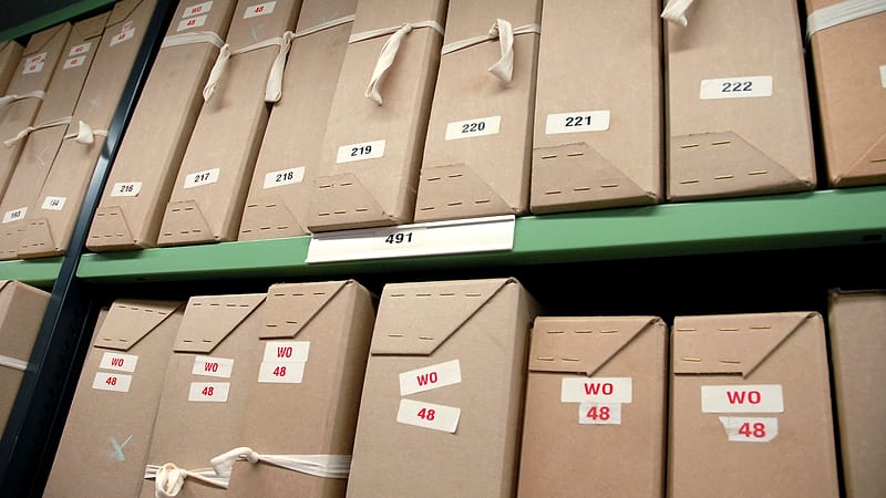 Stacked cardboard document boxes lining a hallway wall, each sealed and labeled with a blank white card.