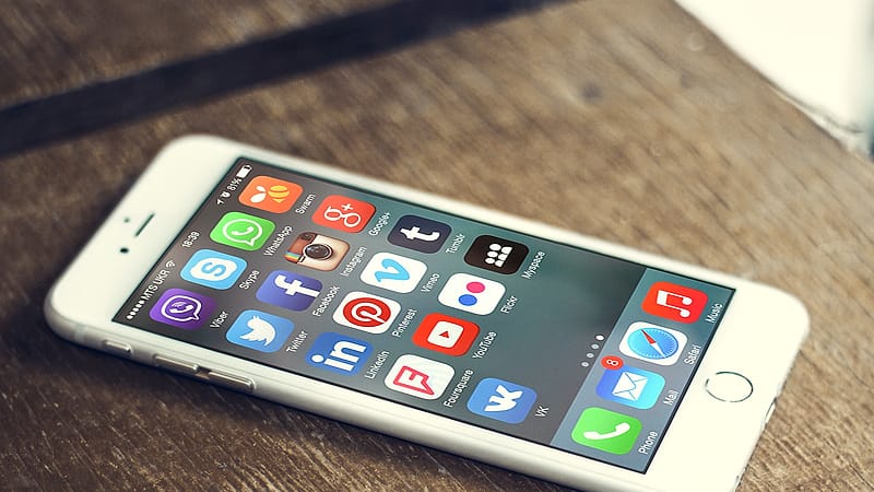 A phone resting face up on a wooden desk beside a passport and a printed form, screen showing a list of app icons.