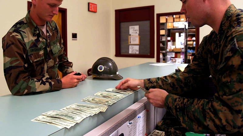 A public service counter with a numbered ticket display, a monitor turned toward the desk, and a folder of copied documents.