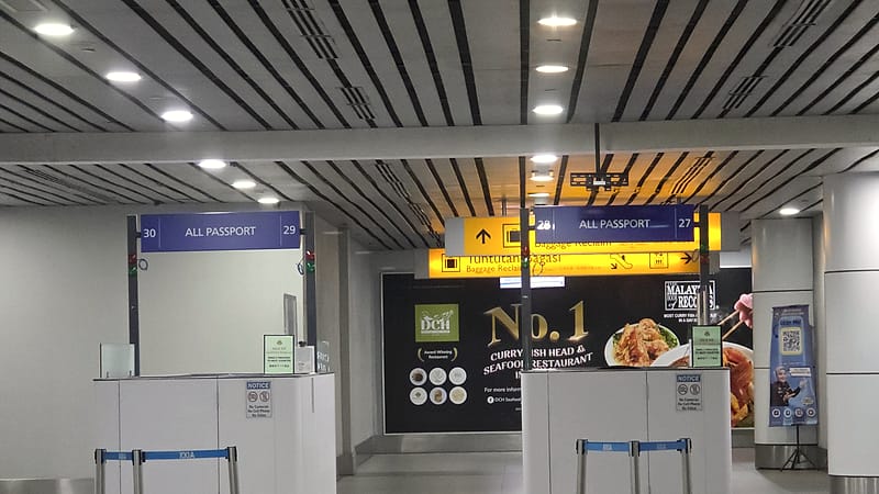 An arrivals hall with a snaking queue of travelers, overhead signage, and rows of inspection booths ahead.