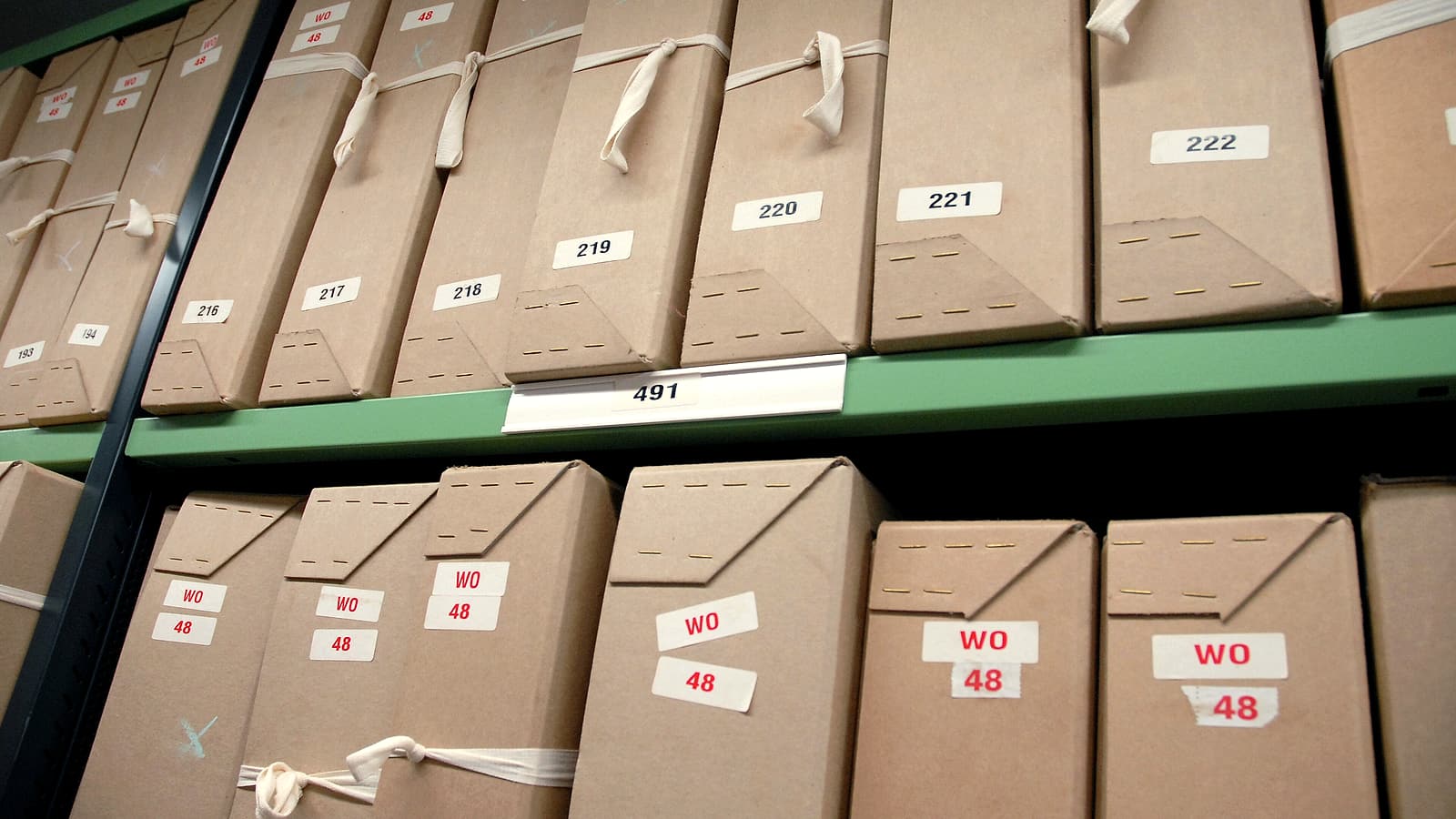 Stacked cardboard document boxes lining a hallway wall, each sealed and labeled with a blank white card.